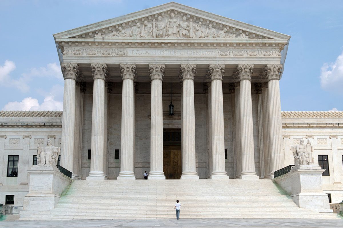 The front facade of the United States Supreme Court building with tall columns, a wide staircase, and two people standing on the steps.