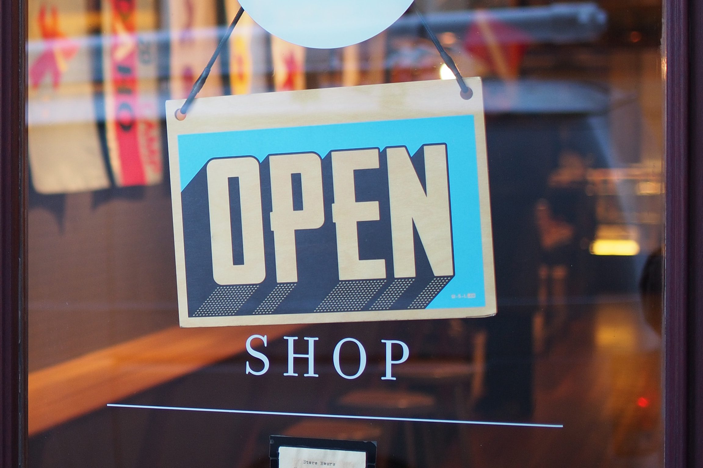 A blue and yellow OPEN sign hangs in the window above the word SHOP on a glass door, indicating the store is open for business.