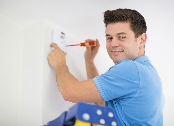 A man in a blue shirt uses a screwdriver to install or adjust a wall-mounted device, possibly a smoke detector, while standing on a ladder.
