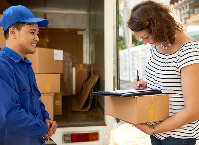 A delivery person in blue uniform watches a woman sign for a package while standing next to a delivery truck with boxes inside.