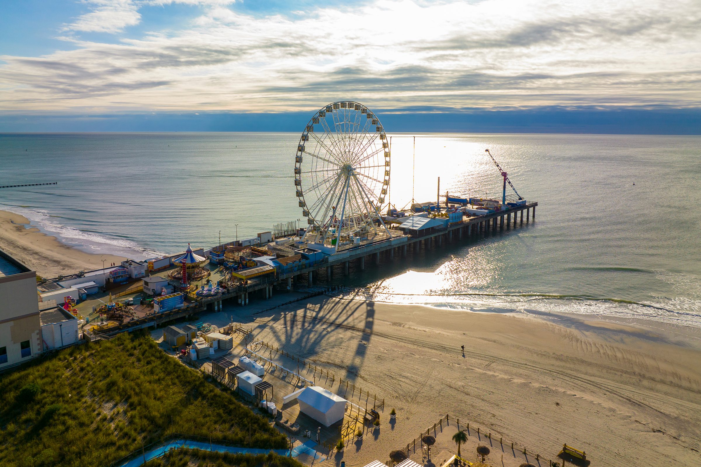 A large Ferris wheel stands on a pier extending into the ocean, with an empty sandy beach and scattered clouds in the sky.
