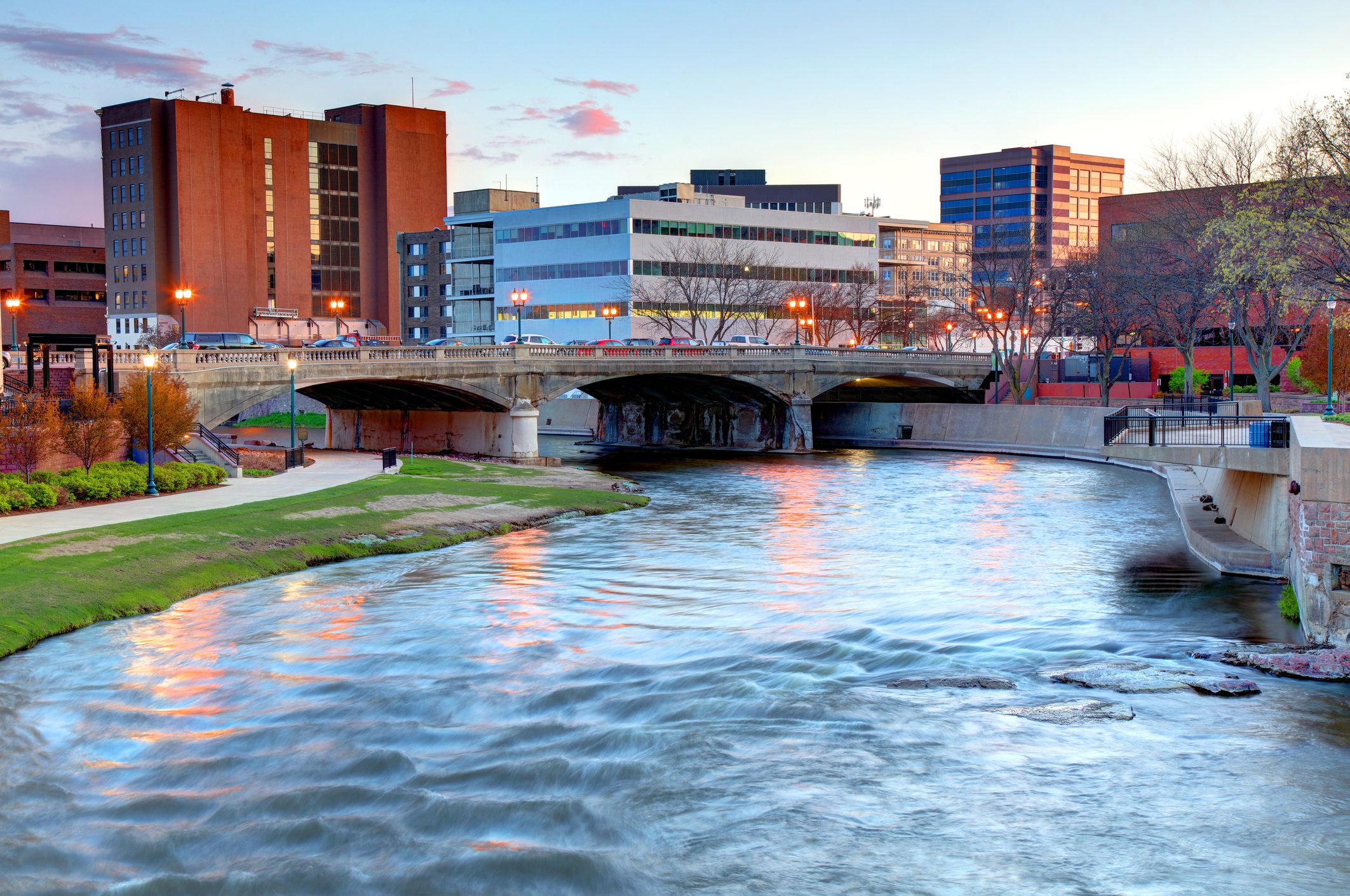 A river flows under a concrete bridge in an urban area with multi-story buildings at sunset.