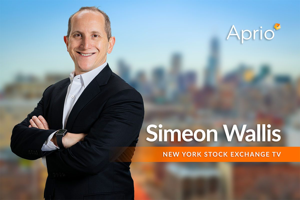 A man in a suit stands smiling with arms crossed in front of a cityscape background. Text reads: Simeon Wallis, New York Stock Exchange TV, Aprio.