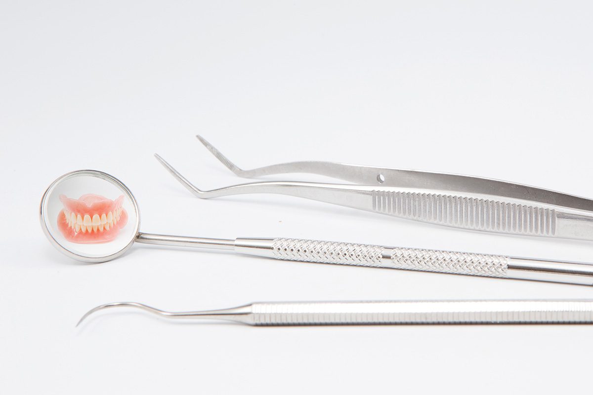 Three dental tools are arranged on a white surface; the mirror tool reflects a set of dentures.