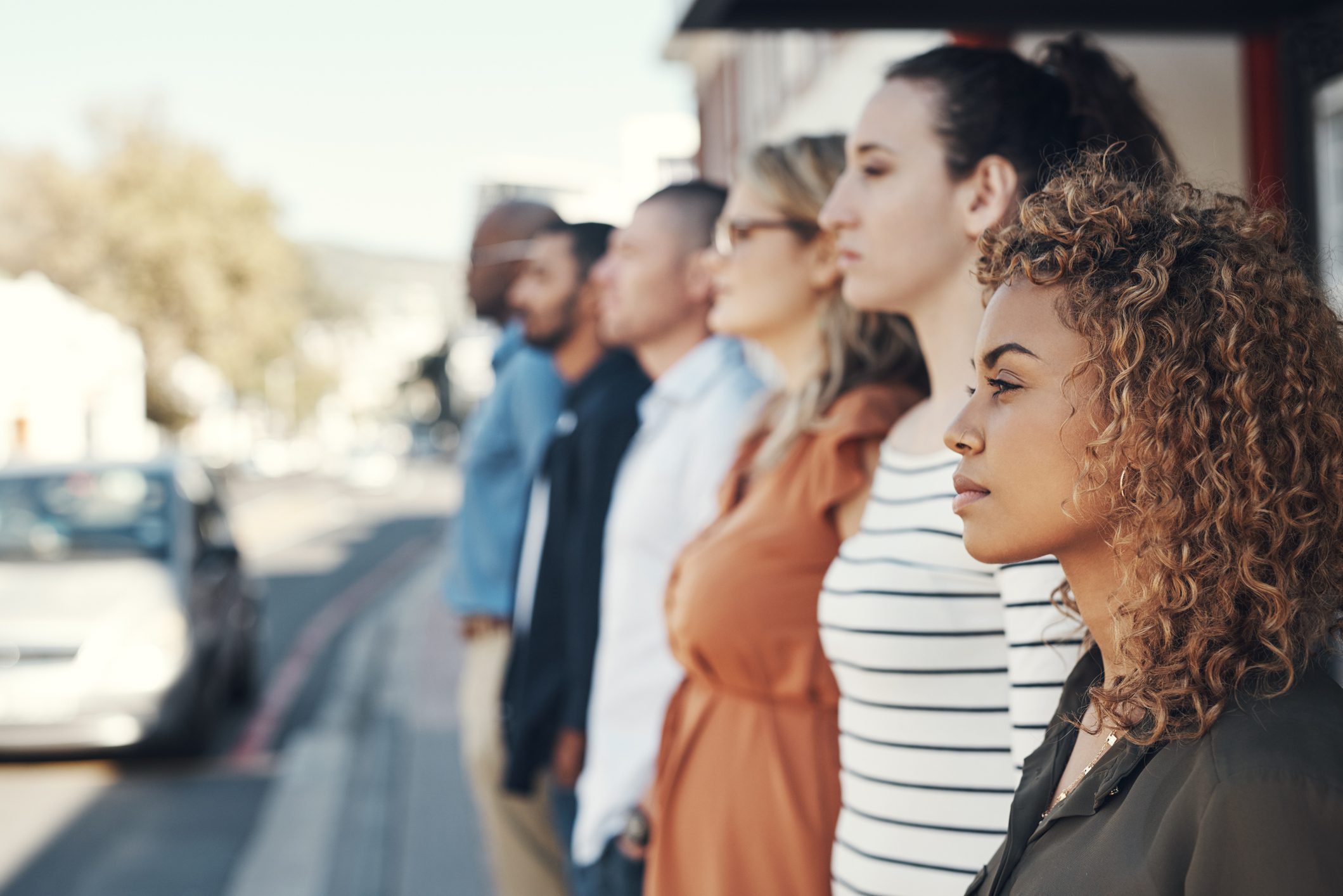 A group of six people stand in a row outdoors on a sunny day, looking in the same direction toward the street.