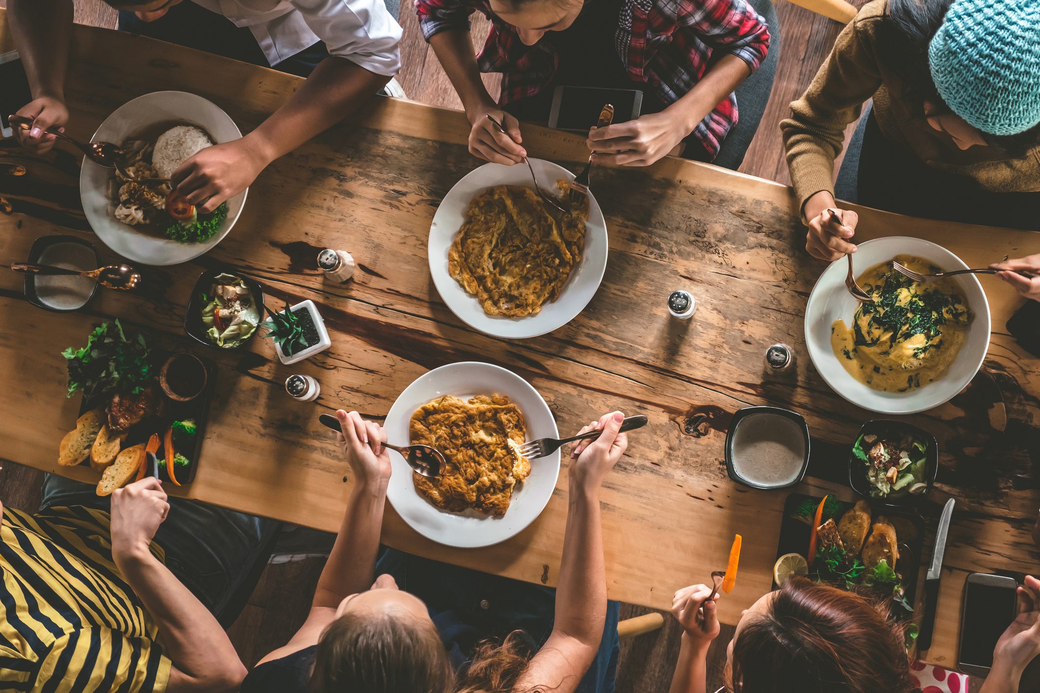 Shot from above, friends sit at a long wooden table eating giant bowls of food