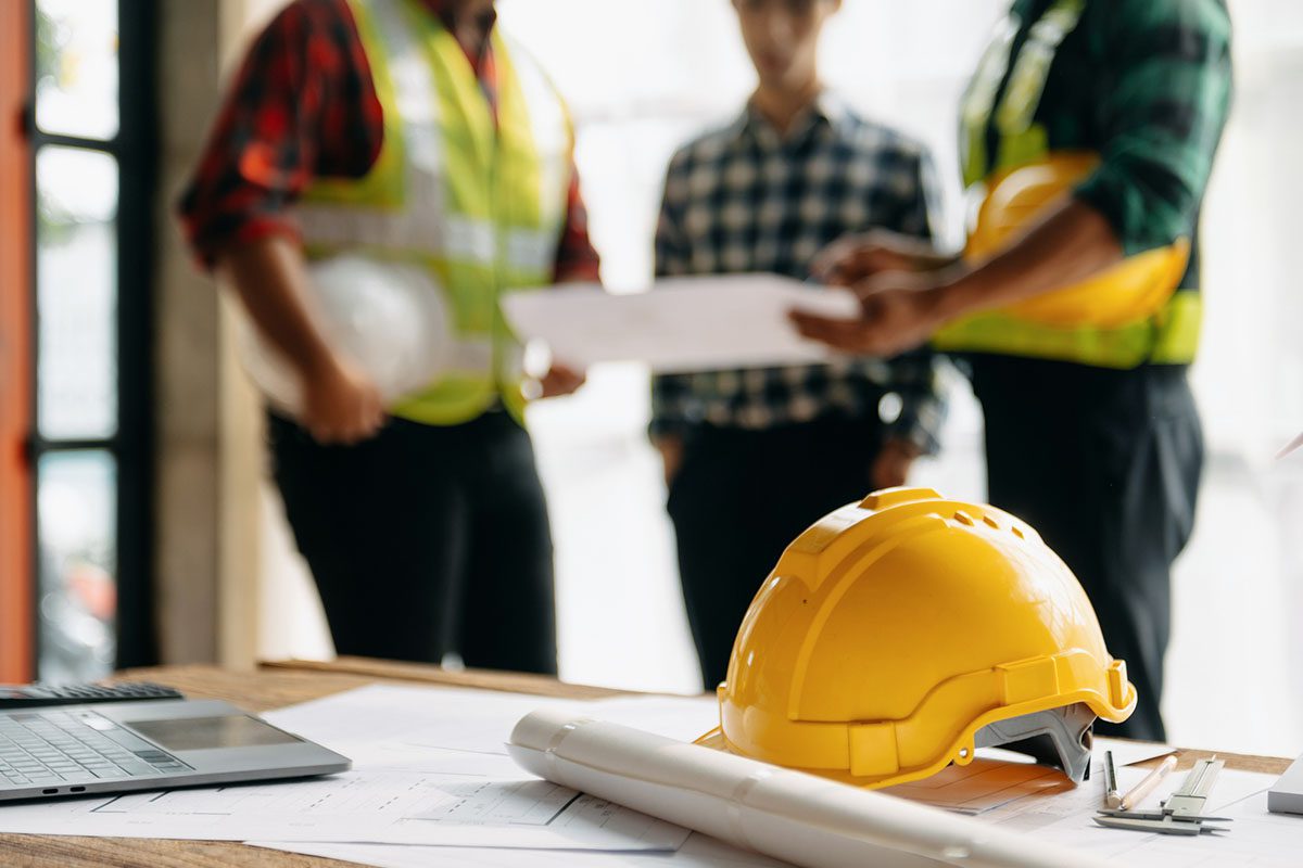 A yellow hard hat, blueprints, and a laptop on a table with three people in safety vests discussing documents in the background.