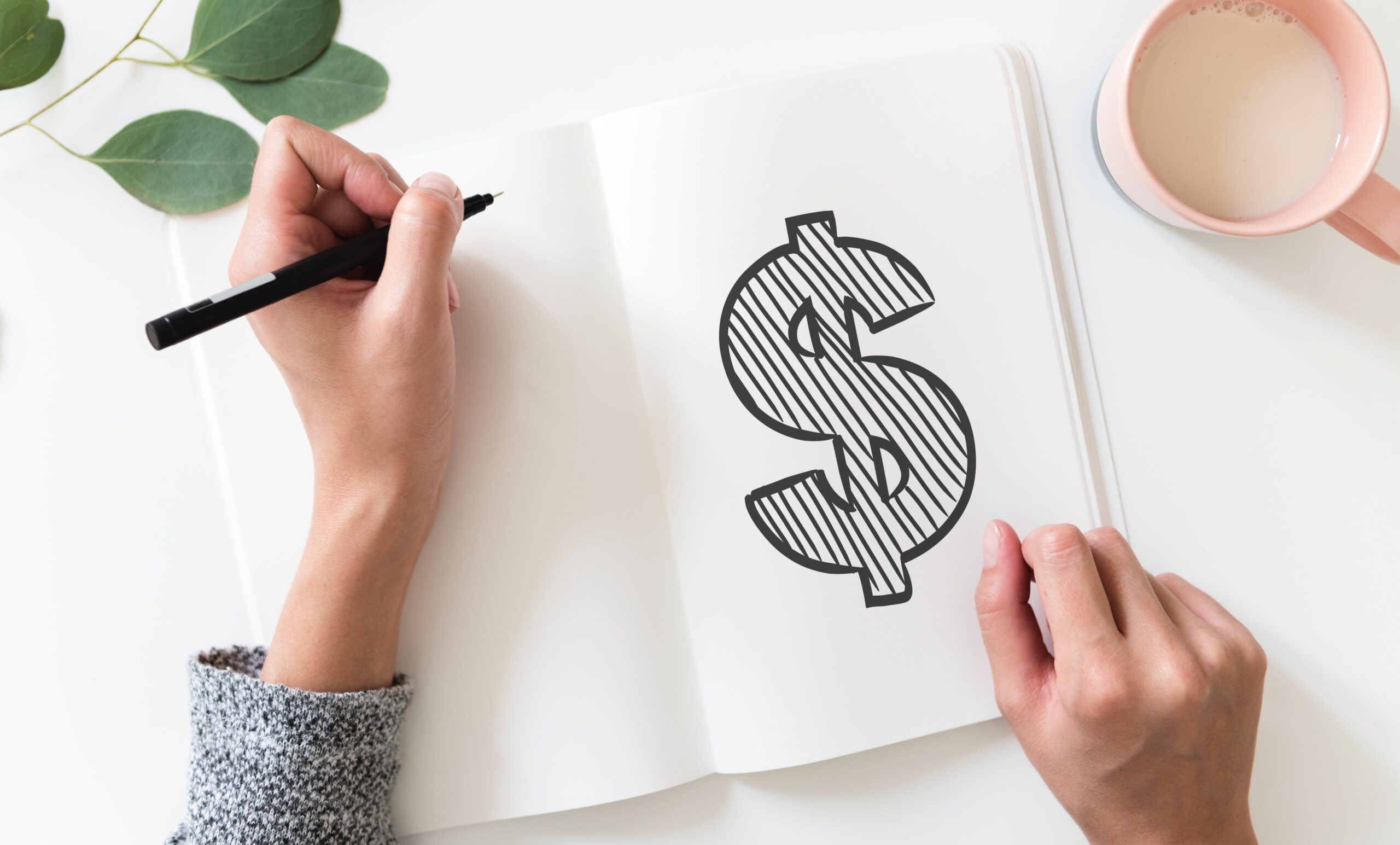 A person holds a pen and draws a large dollar sign in a notebook, with a pink coffee cup and green leaves nearby on a white surface.
