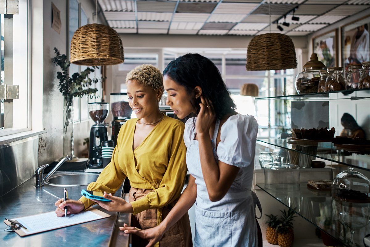 Two women stand at a counter in a cafe or bakery, reviewing paperwork together; one holds a pen and the other looks at a digital tablet.