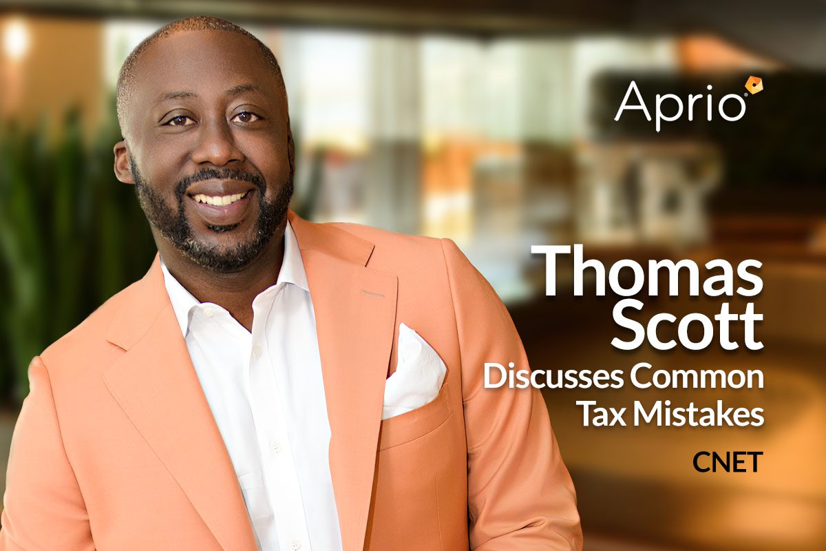 A man in a peach suit jacket smiles at the camera. Text reads: Aprio. Thomas Scott Discusses Common Tax Mistakes. CNET. The background is a blurred office space.
