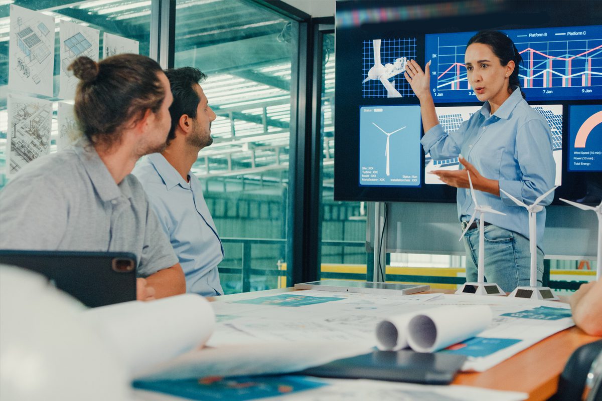 A woman presents renewable energy data on a screen to two colleagues seated at a table with architectural plans and wind turbine models.