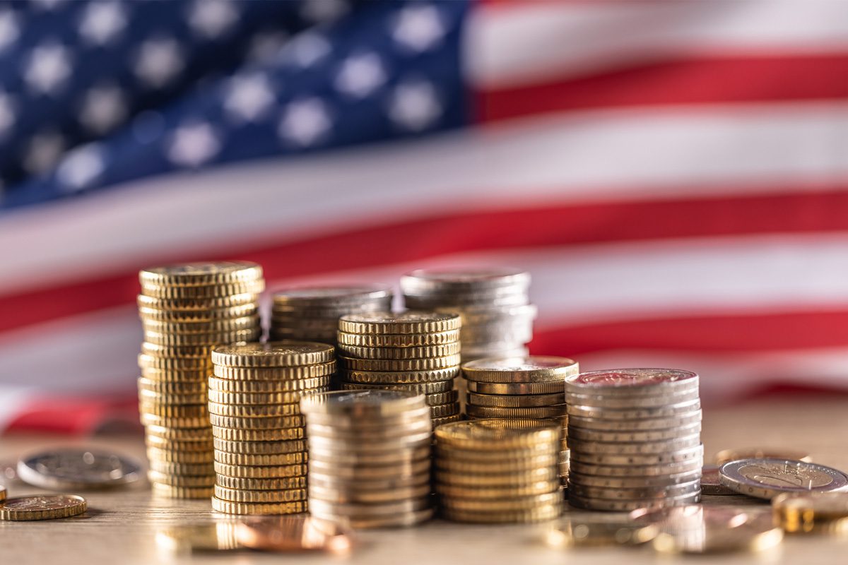 Stacks of assorted coins are arranged on a table with a blurred American flag in the background.