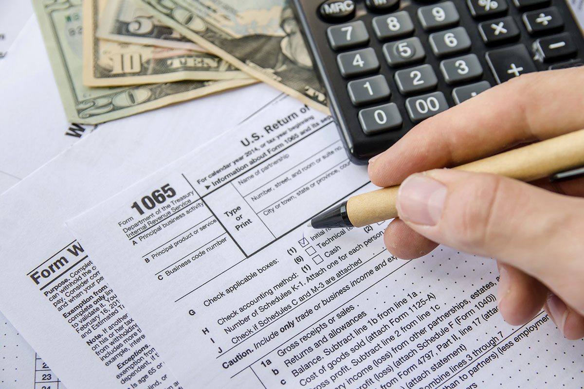 A person fills out a tax form 1065 with a pen, alongside a calculator, cash, and other documents on a desk.