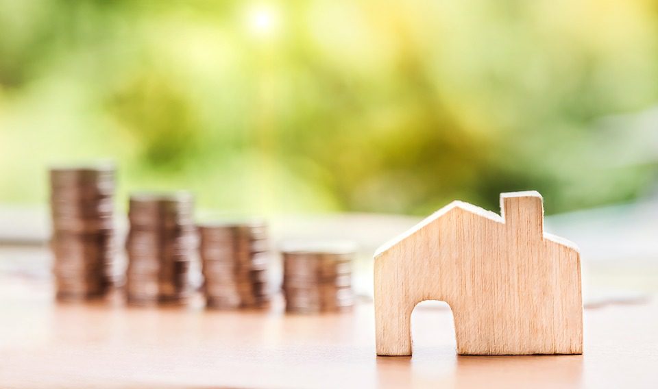 Stacks of coins gradually increasing in height are placed next to a small wooden house model on a table, with a blurred natural background.