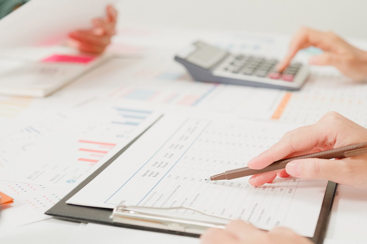 Two people are working with financial documents, graphs, and a calculator on a desk. One person writes on a clipboard while the other uses the calculator.