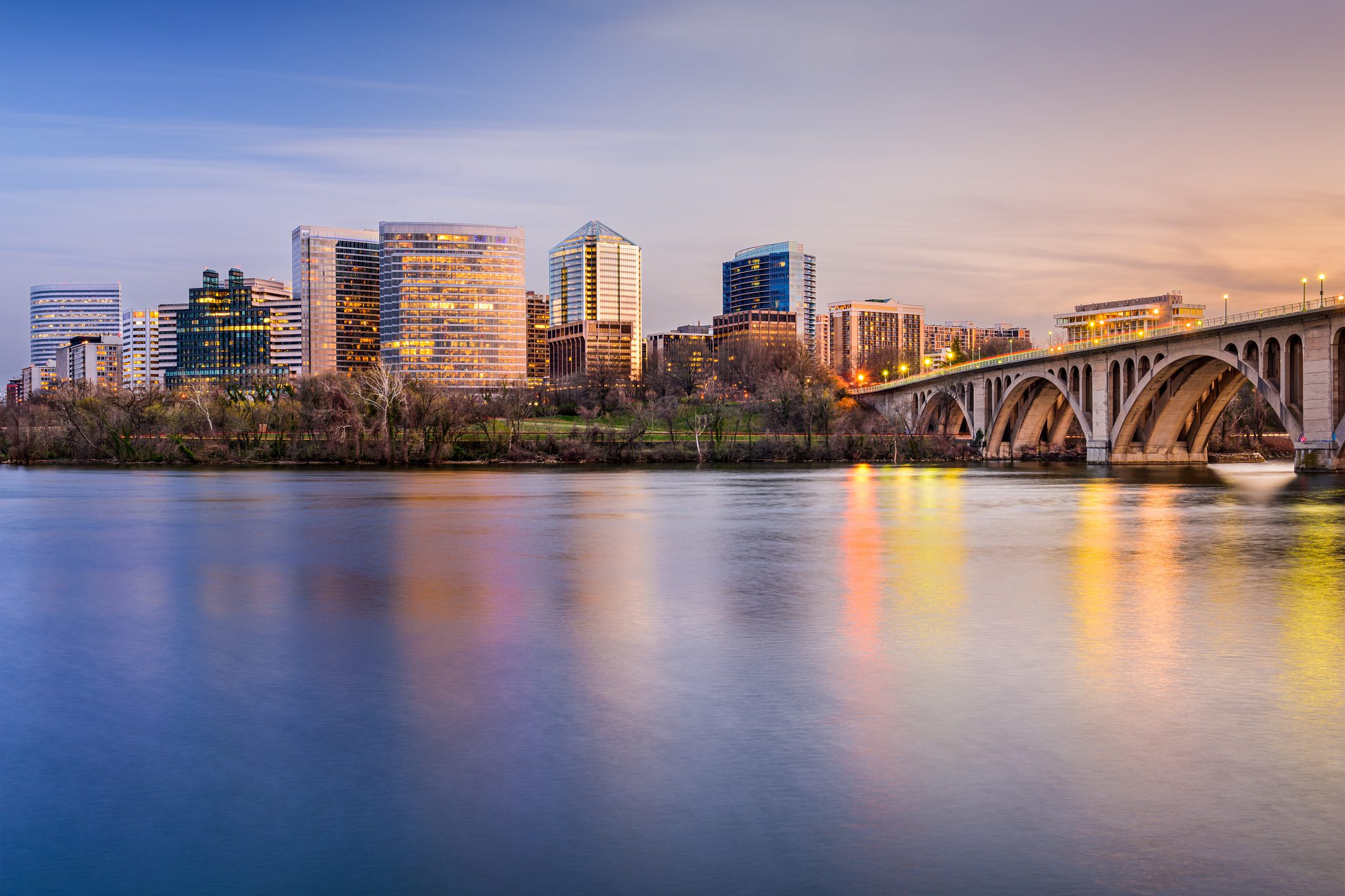 City skyline with modern high-rise buildings and a large arched bridge reflected in a calm river at dusk.