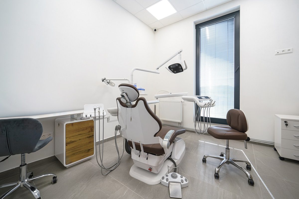Modern dental examination room with a dental chair, overhead light, instruments, two stools, and a large window with blinds.