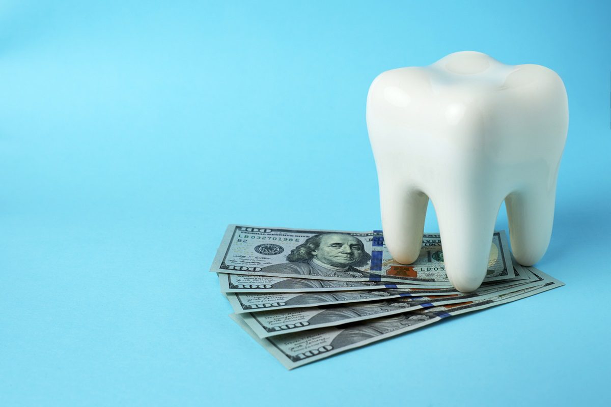 A large model of a tooth is placed on top of several hundred-dollar bills against a blue background.