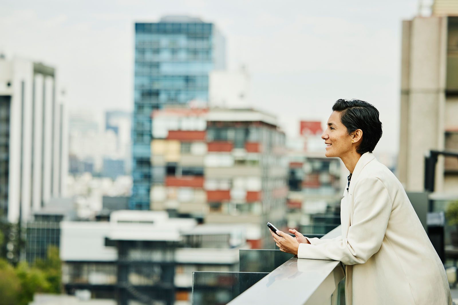 Person in a white blazer stands on a balcony, holding a smartphone, looking out over a modern cityscape with tall buildings in the background.