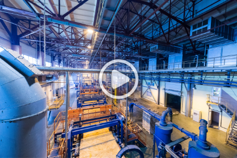 Interior view of an industrial facility with large machinery and overhead metal beams, lit by bright blue and yellow lighting, with a play button icon in the center.