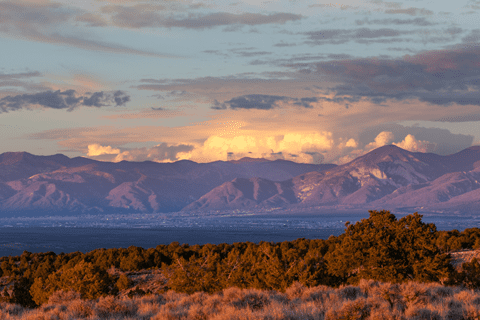 Mountains under a partly cloudy sky at sunset, with sunlight casting a warm glow on the landscape and trees in the foreground.
