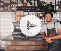 Man wearing an apron and hat stands smiling behind a wooden counter in a modern café with shelves and plants in the background.