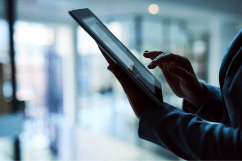 Person using a touchscreen tablet indoors, with blurred background and natural light coming through windows.